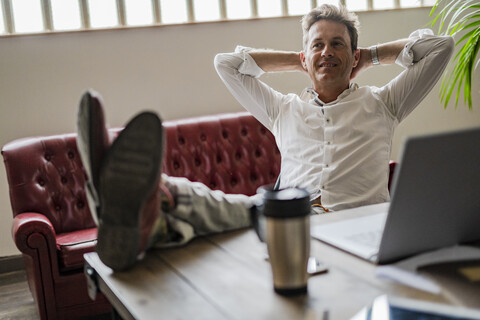 Lächelnder Geschäftsmann sitzt mit den Füßen auf dem Schreibtisch, lizenzfreies Stockfoto