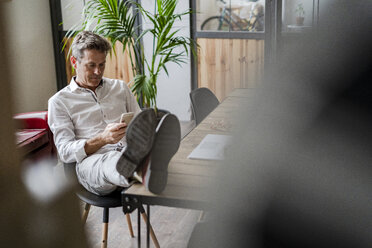 Businessman sitting with feet on desk using cell phone - GIOF05069