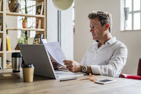 Konzentrierter Geschäftsmann, der einen Laptop benutzt und Dokumente am Schreibtisch durchgeht - GIOF05074