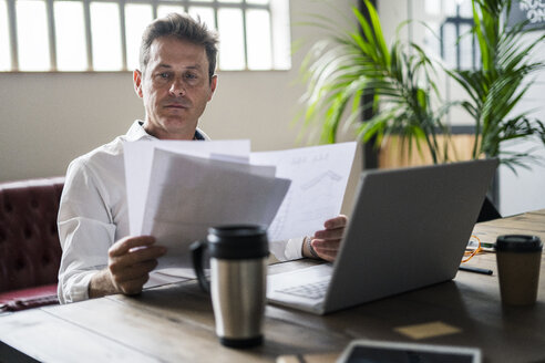 Konzentrierter Geschäftsmann, der einen Laptop benutzt und Dokumente am Schreibtisch durchgeht - GIOF05075