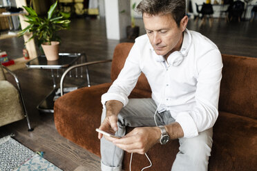 Mature man with cell phone and headphones sitting on couch in a loft - GIOF05089