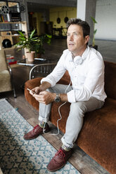 Mature man with cell phone and headphones sitting on couch in a loft thinking - GIOF05090