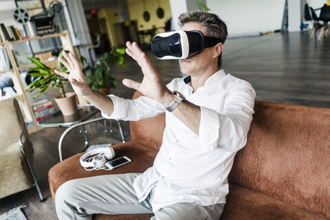 Älterer Mann mit VR-Brille sitzt auf einer Couch in einem Loft, lizenzfreies Stockfoto