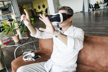 Mature man wearing VR glasses sitting on couch in a loft - GIOF05096