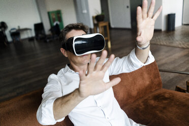 Mature man wearing VR glasses sitting on couch in a loft - GIOF05098