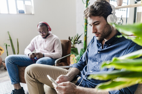 Zwei junge Männer mit Handys und Kopfhörern sitzen in Sesseln in einem Loft, lizenzfreies Stockfoto