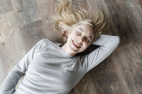 Happy blond young woman lying on wooden floor - GIOF05139