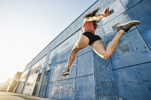 Niedriger Blickwinkel auf eine Sportlerin, die auf dem Bürgersteig an einem blauen, mit Graffiti beschmierten Gebäude vorbeiläuft., lizenzfreies Stockfoto