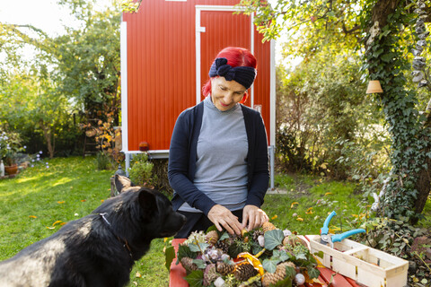 Lächelnde ältere Frau mit rot gefärbtem Haar und Hund beim Basteln von Herbstdekoration im Garten, lizenzfreies Stockfoto