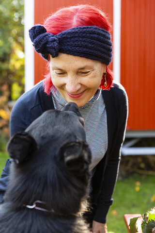 Porträt einer lächelnden älteren Frau mit rot gefärbtem Haar von Angesicht zu Angesicht mit ihrem Hund im Garten, lizenzfreies Stockfoto