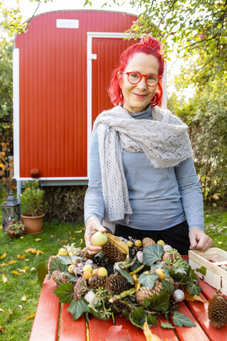 Porträt einer lächelnden älteren Frau mit rot gefärbten Haaren, die im Garten an einer herbstlichen Dekoration bastelt, lizenzfreies Stockfoto