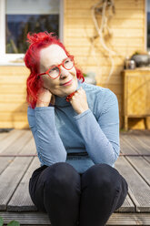 Porträt einer lächelnden älteren Frau mit rot gefärbtem Haar, die auf der Terrasse vor ihrem Haus sitzt und sich entspannt - OJF00310