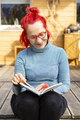 Porträt einer lächelnden älteren Frau mit rot gefärbtem Haar, die auf der Terrasse vor ihrem Haus sitzt und ein Buch liest - OJF00311