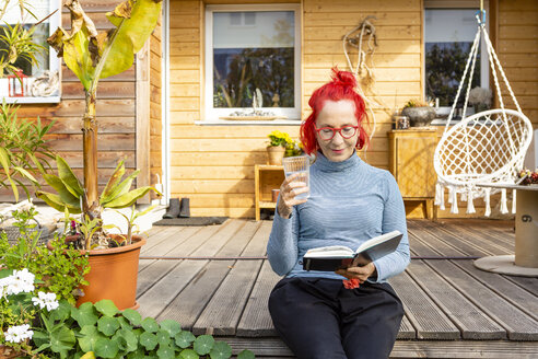 Porträt einer lächelnden älteren Frau mit rot gefärbtem Haar, die auf der Terrasse vor ihrem Haus sitzt und ein Buch liest - OJF00313