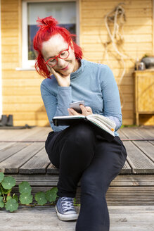Porträt einer lächelnden älteren Frau mit rot gefärbtem Haar, die auf der Terrasse vor ihrem Haus sitzt und ein Handy benutzt - OJF00315