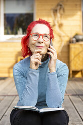 Porträt einer lächelnden älteren Frau mit rot gefärbtem Haar, die auf der Terrasse vor ihrem Haus sitzt und telefoniert - OJF00316