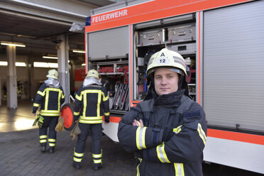 Portrait of confident firefighter in front of fire engine with colleagues in background - LYF00845