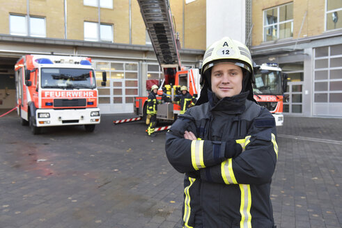 Porträt eines selbstbewussten Feuerwehrmanns vor einem Löschfahrzeug mit Kollegen im Hintergrund - LYF00855