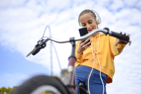 Smiling girl with headphones and bicycle looking at smartphone stock photo