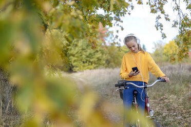 Lächelndes Mädchen mit Kopfhörern, das auf einem Fahrrad sitzt und auf sein Smartphone schaut - BFRF01952