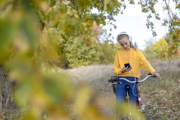 Smiling girl with headphones sitting on bicycle looking at smartphone - BFRF01952