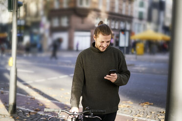 Lächelnder junger Mann mit Fahrrad in der Stadt schaut auf sein Handy - FMKF05337