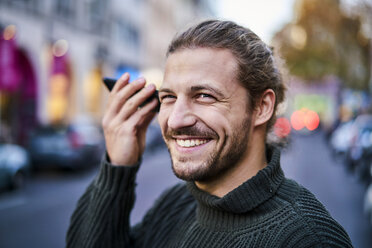 Porträt eines lachenden jungen Mannes, der abends auf der Straße sein Smartphone benutzt - FMKF05346