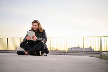 Porträt eines bärtigen jungen Mannes, der in der Dämmerung auf einem Parkdeck sitzt und ein digitales Tablet benutzt - FMKF05349