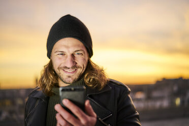 Porträt eines bärtigen jungen Mannes mit Wollmütze, der bei Sonnenuntergang auf sein Mobiltelefon schaut - FMKF05350