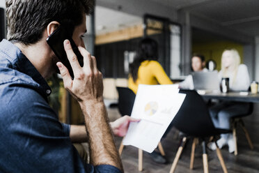 Geschäftsmann, der Dokumente in der Hand hält und während einer Besprechung im Büro mit seinem Handy telefoniert - GIOF05195