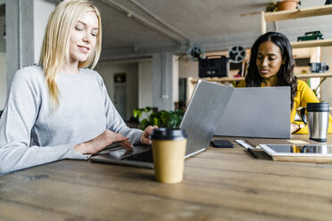 Zwei junge Geschäftsfrauen sitzen am Konferenztisch in einem Loft-Büro und benutzen Laptops - GIOF05205