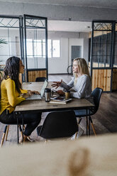 Two young businesswomen talking at conference table in loft office - GIOF05210