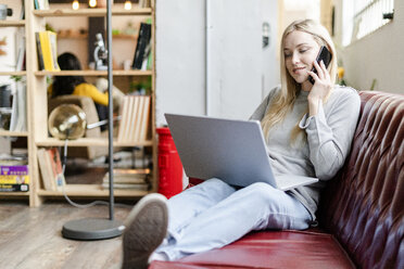 Lächelnde junge Frau sitzt mit Laptop auf der Couch und spricht mit dem Handy - GIOF05225