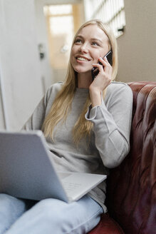 Lächelnde junge Frau sitzt mit Laptop auf der Couch und spricht mit dem Handy - GIOF05228
