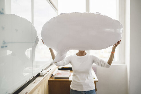 Frau steht im Büro und hält sich eine aufblasbare Sprechblase vor das Gesicht - RIBF00774