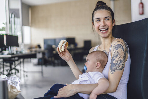 Junge Mutter mit Tattoos sitzt im Büro mit ihrem Baby auf dem Schoß und isst ein Sandwich - RIBF00777