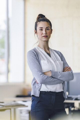 Porträt einer schönen jungen Frau, stehend im Büro, mit verschränkten Armen, lizenzfreies Stockfoto