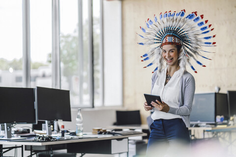 Junge Frau mit indianischem Kopfschmuck, im Büro stehend, mit digitalem Tablet, lizenzfreies Stockfoto