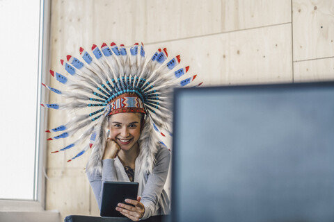 Junge Frau mit indianischem Kopfschmuck, im Büro stehend, mit digitalem Tablet, lizenzfreies Stockfoto