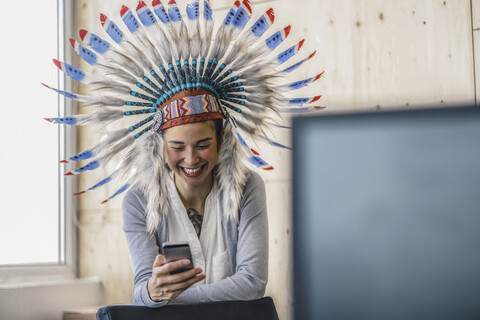 Junge Frau mit indianischem Kopfschmuck, steht im Büro und benutzt ein Smartphone, lizenzfreies Stockfoto