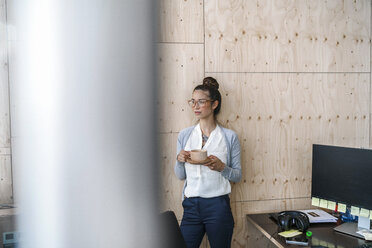 Junge Frau arbeitet in einem kreativen Büro, macht eine Pause und trinkt Kaffee aus einer Holztasse - RIBF00795