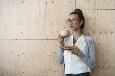 Junge Frau arbeitet in einem kreativen Büro, macht eine Pause und trinkt Kaffee aus einer Holztasse - RIBF00796