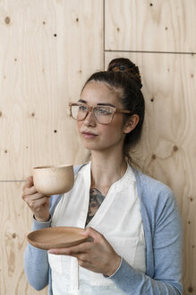 Junge Frau arbeitet in einem kreativen Büro, macht eine Pause und trinkt Kaffee aus einer Holztasse - RIBF00797