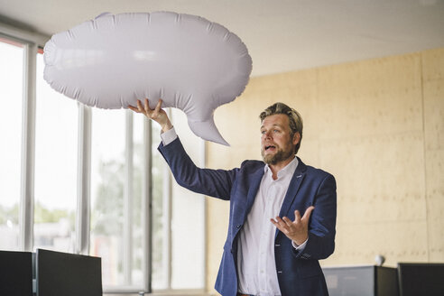 Businessman standing in office, holding inflatable speech bubble - RIBF00798