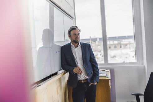 Geschäftsmann steht in einem Büro, hält eine Brille in der Hand und stützt sich auf eine weiße Tafel - RIBF00806