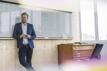 Businessman standing in office, holding smartphone, leaning on white board - RIBF00808