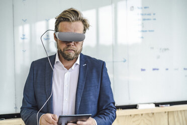 Businessman standing in office, using VR glasses and digital tablet - RIBF00809