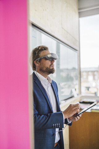 Geschäftsmann im Büro stehend, mit VR-Brille und digitalem Tablet, lizenzfreies Stockfoto