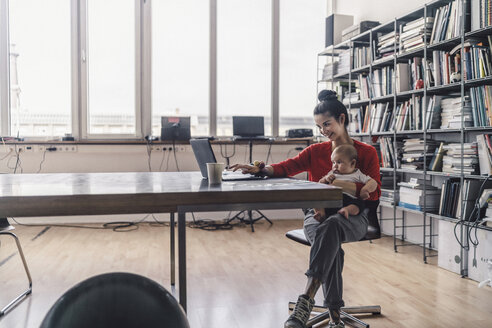 Arbeitende Mutter mit Baby auf dem Schoß, im Büro sitzend, mit Laptop - RIBF00822