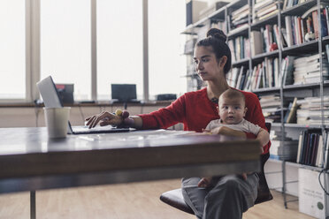 Arbeitende Mutter mit Baby auf dem Schoß, im Büro sitzend, mit Laptop - RIBF00823